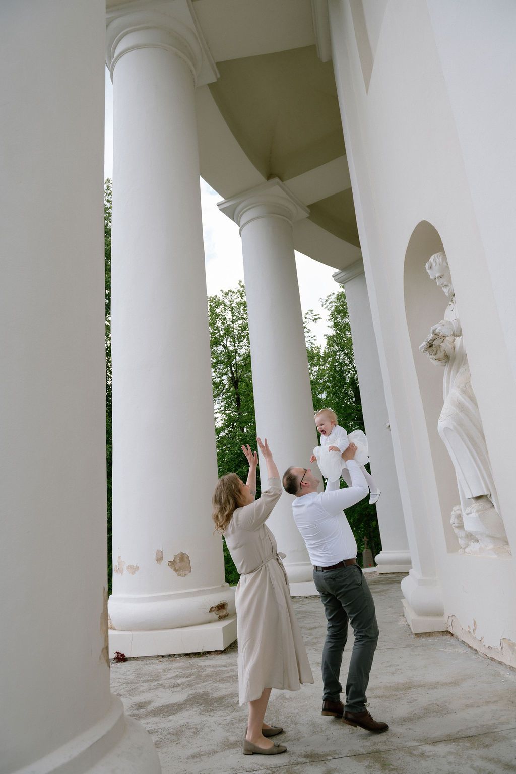 Family with baby enjoying a classical white colonnade with elegant statues and lush green trees in the background.