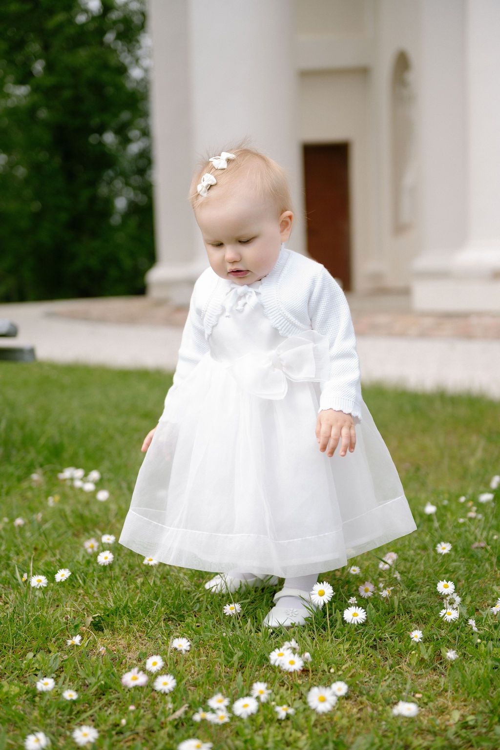 A baby girl in a white dress and flower headband stands on green grass with daisies outside a white building.