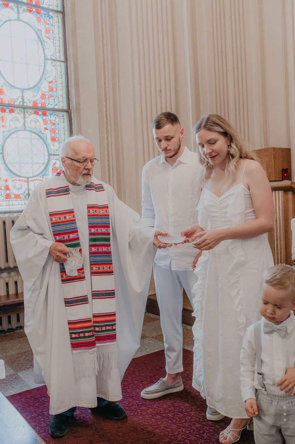 A priest officiates a wedding ceremony with a couple in white attire and a young child in a church.