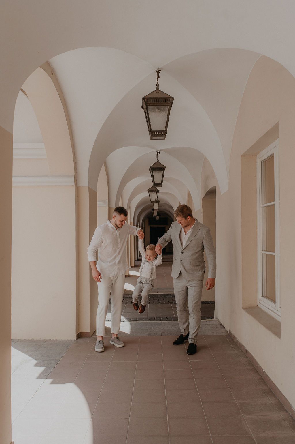 Two men in light suits swing a toddler between them in a sunlit arched hallway.