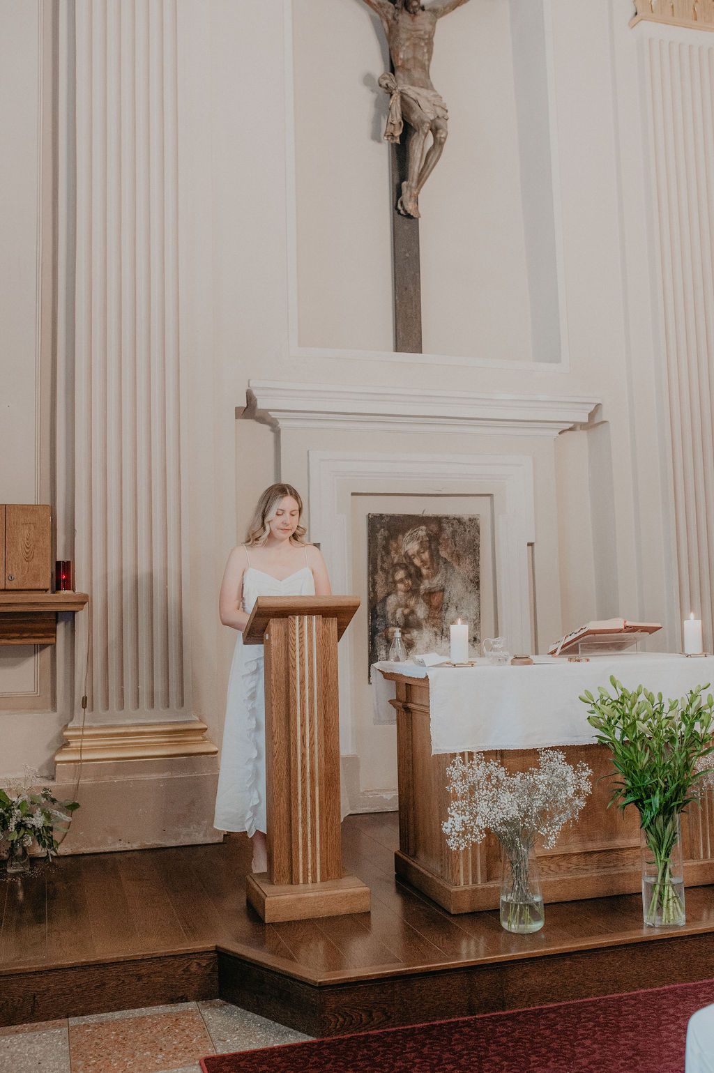 Woman in white dress reading at wooden church pulpit with crucifix and altar in background.
