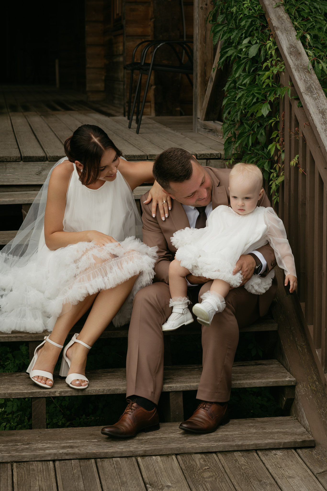 Bride and groom in wedding attire sit on wooden steps holding their baby dressed in white.