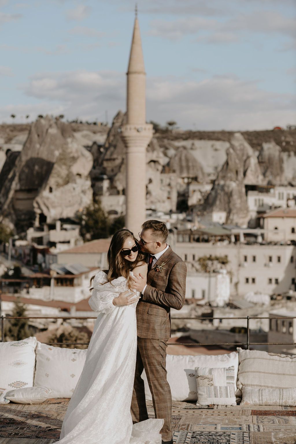 Couple embracing in wedding attire on a rooftop with Cappadocia cave dwellings and mosque minaret backdrop.