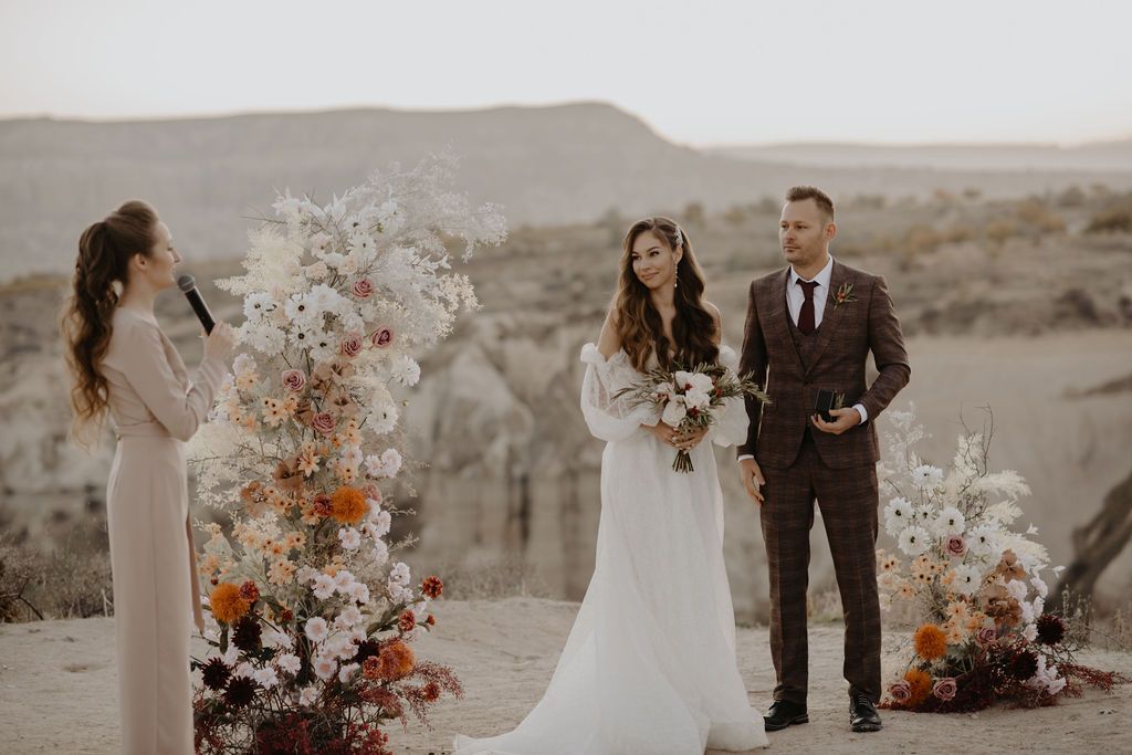 Outdoor desert wedding ceremony with bride holding bouquet, groom in suit, and officiant beside floral arch.