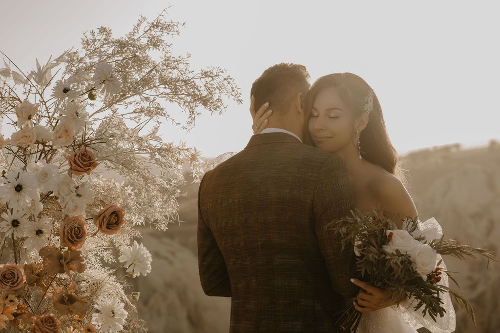 Bride and groom embrace at golden hour with boho floral arrangement in warm tones during outdoor wedding ceremony.