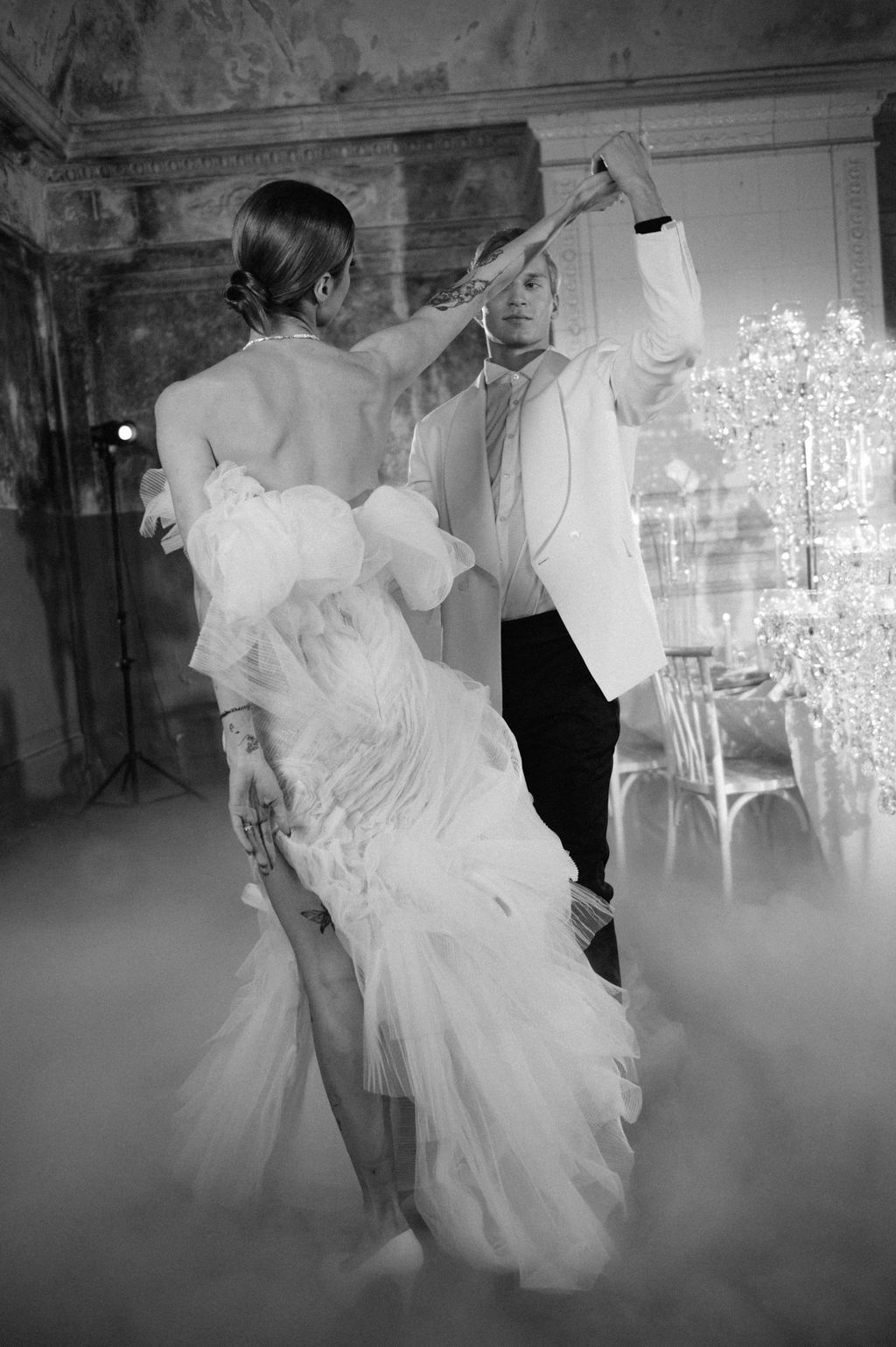 Black and white photo of a couple sharing their first dance, bride in ruffled gown, groom in white jacket, surrounded by mist.