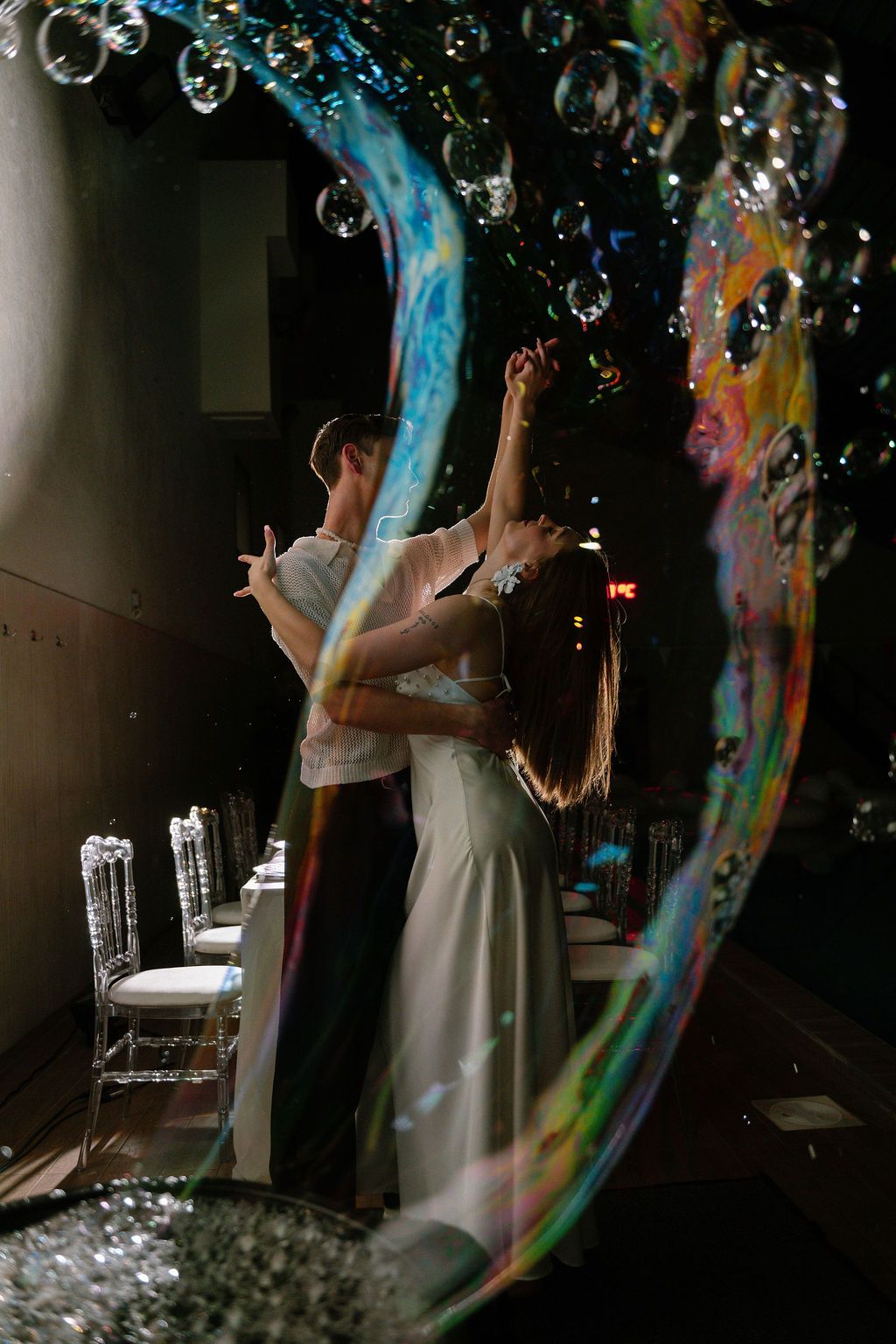 Bride and groom share first dance surrounded by colorful soap bubbles in elegant dark wedding venue.