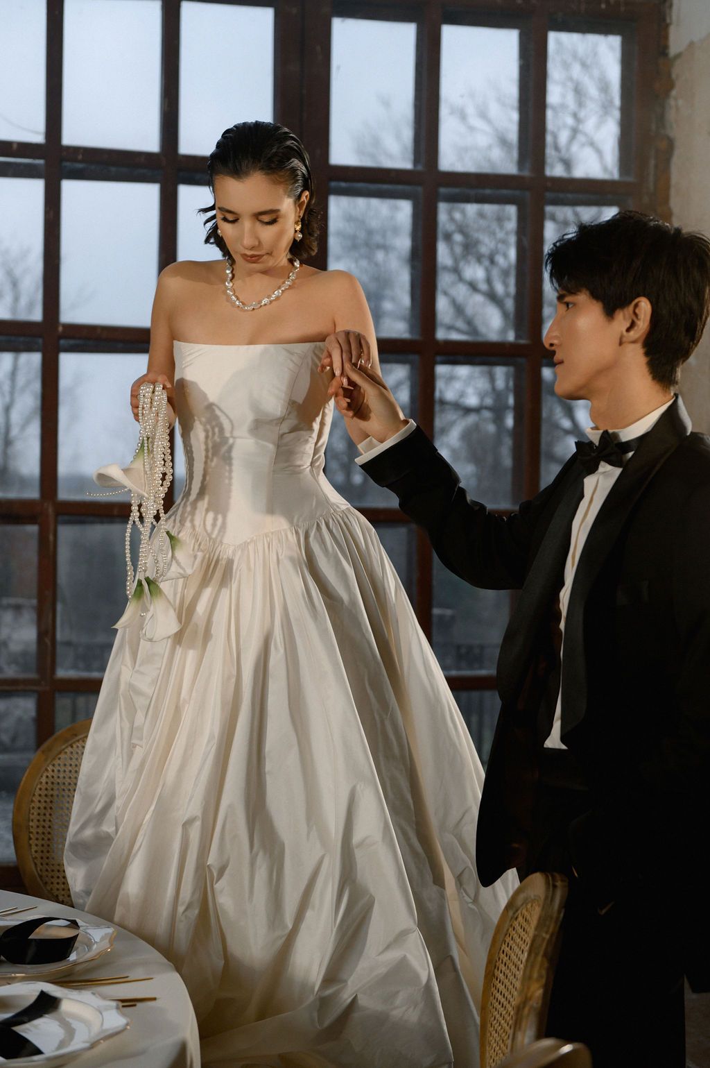Groom in black tuxedo fastening pearl necklace on bride in white strapless ballgown near large windows.