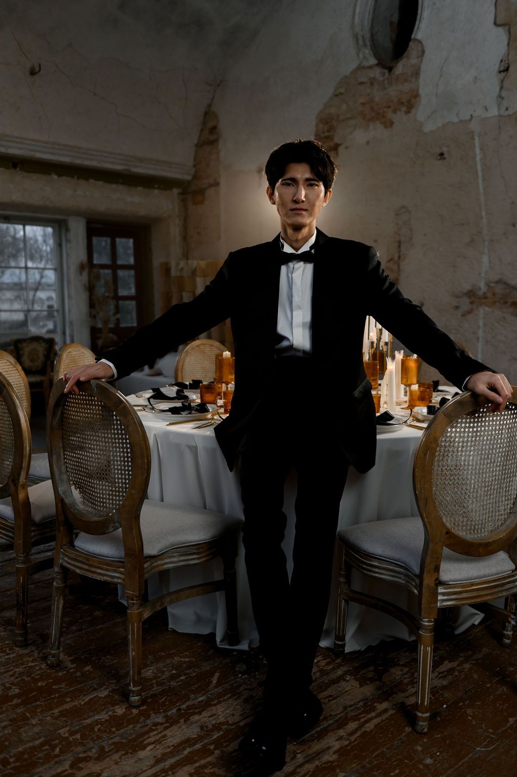 Elegant man in black tuxedo leaning on dining table with ornate chairs.