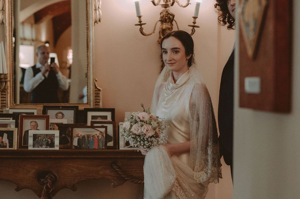 Elegant bride in vintage lace gown holding bouquet stands by antique mirror with framed photos at wedding venue.