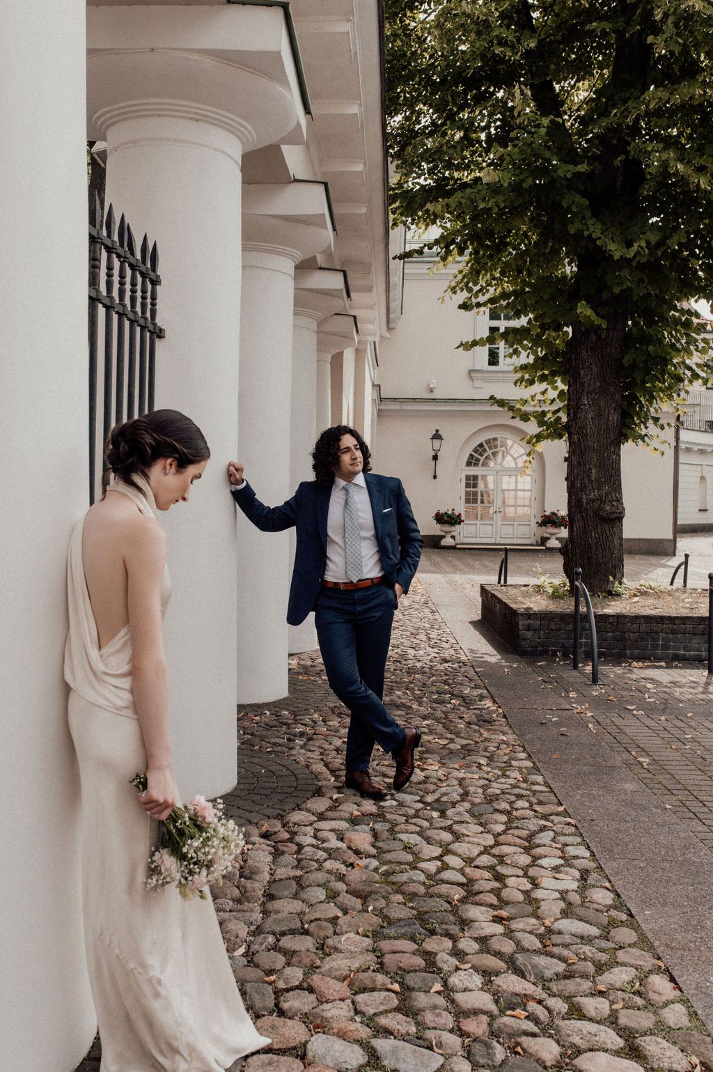 Bride in a satin halter dress and groom in a navy suit share a candid moment by white columns on a cobblestone path.