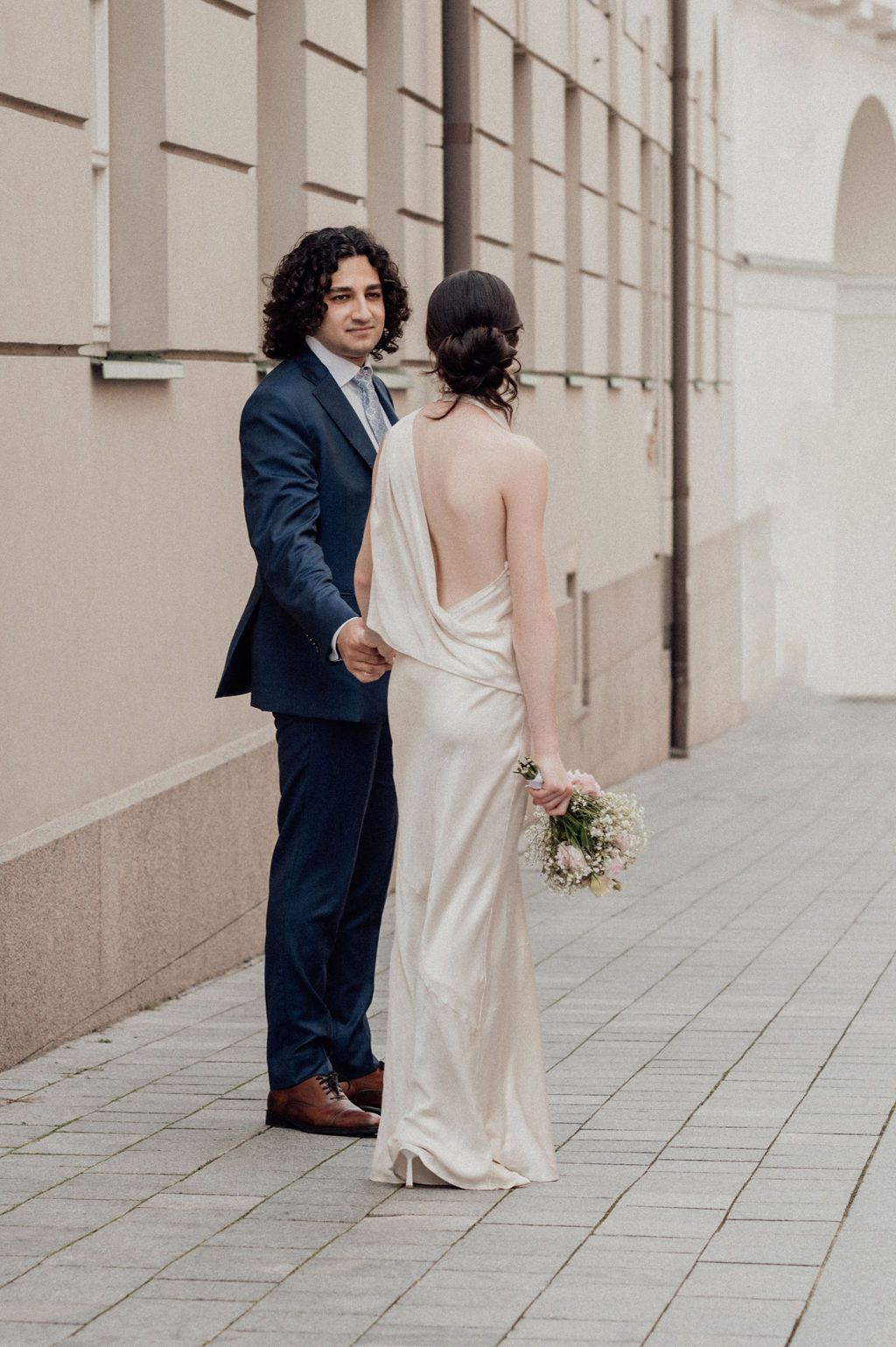 A groom in a navy suit holds hands with a bride in a backless ivory gown, holding a bouquet on a cobblestone street.