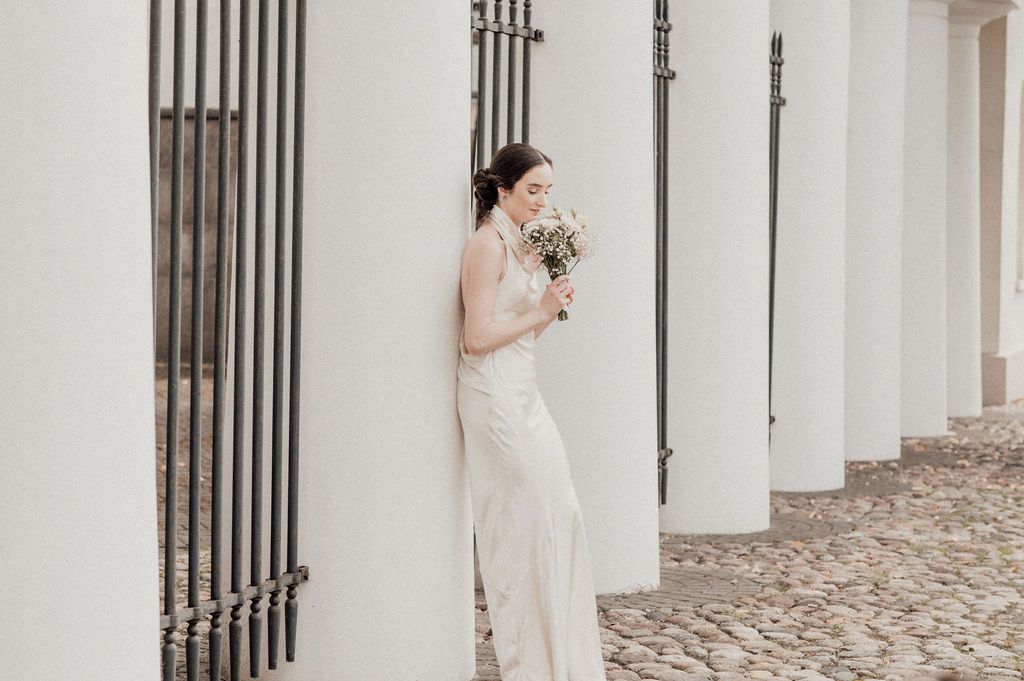 Bride in a white lace wedding dress leaning against a column holding a bouquet on cobblestone.