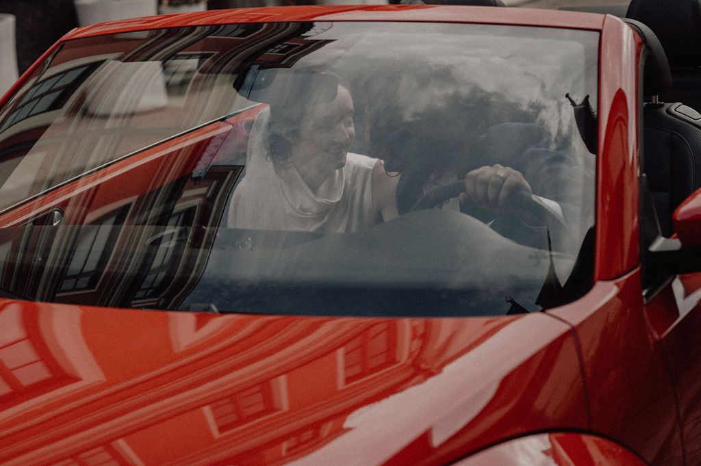 Person smiling inside a red sports car, viewed through the windshield on a sunny day.