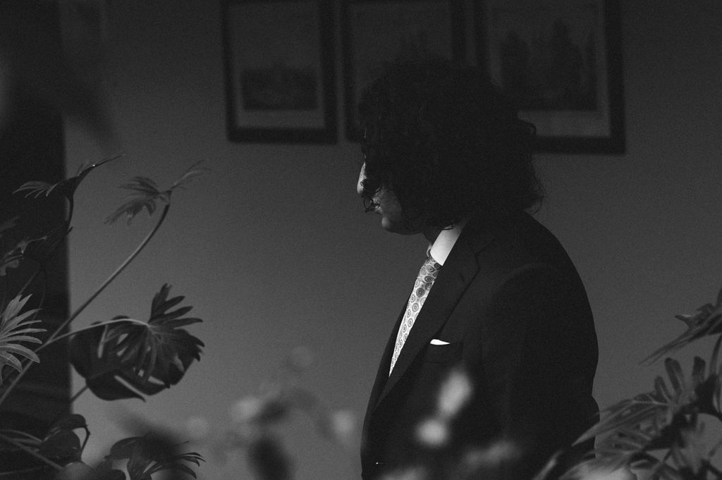 Black and white portrait of a man in a suit with curly hair standing among plants with framed art on wall.