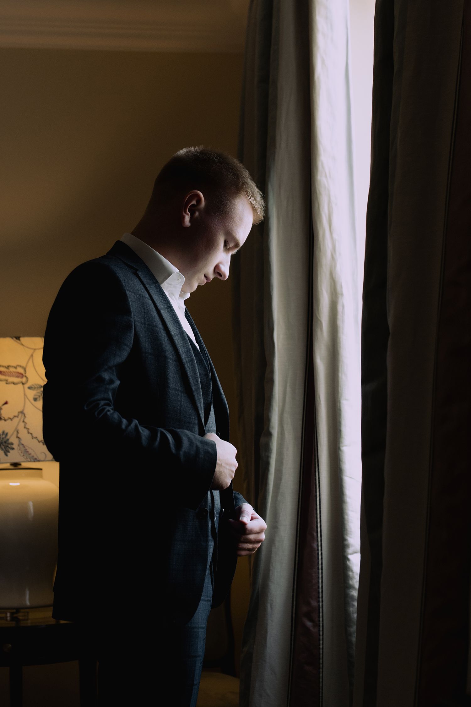 A person in a dark suit stands by a window in dramatic lighting creating a moody silhouette effect.