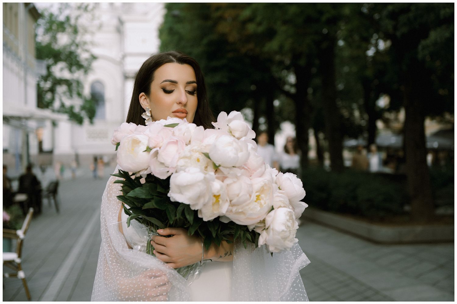 A woman in a white dress holds a lush bouquet of pink and white peonies in an outdoor setting.