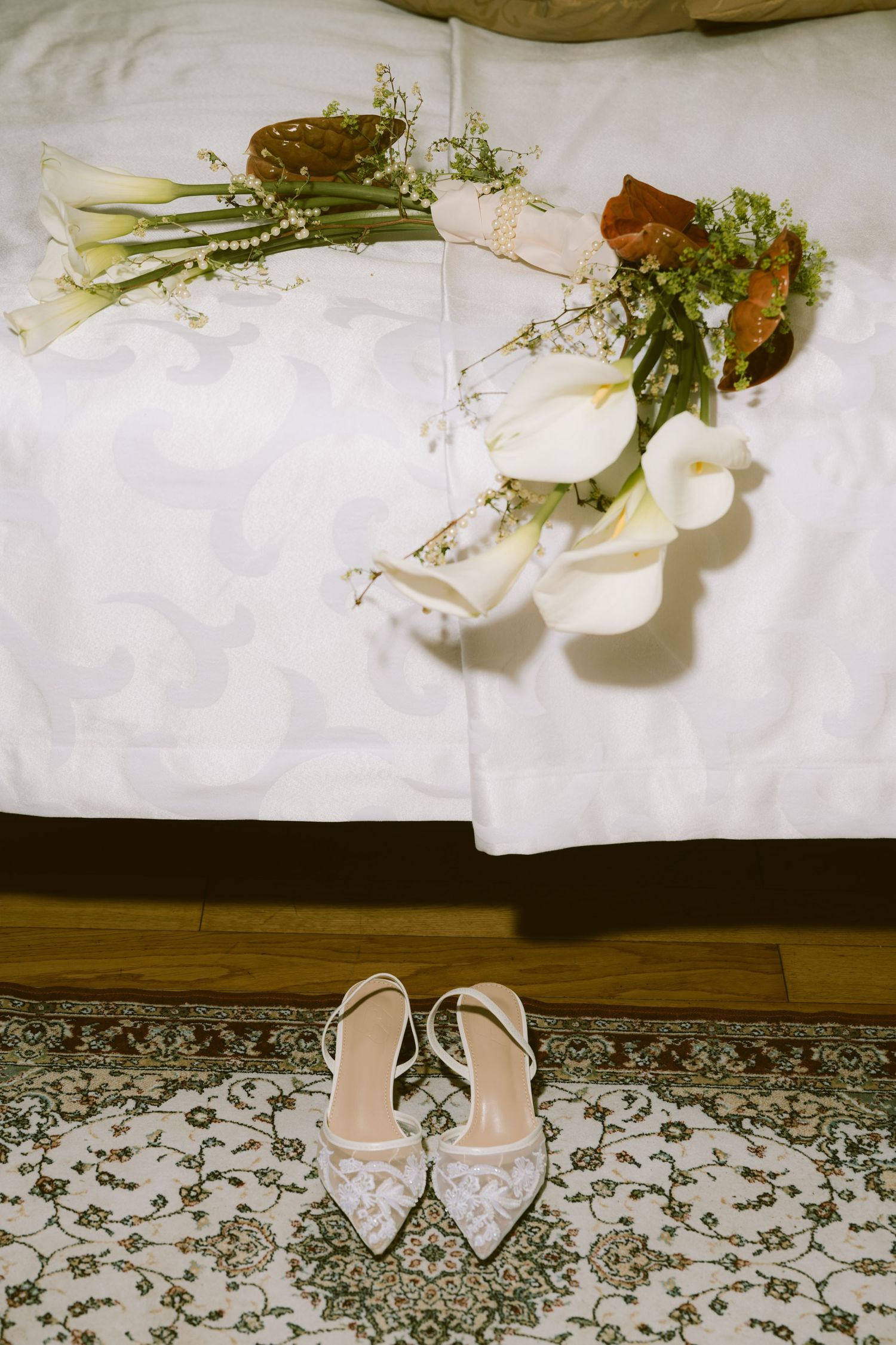 Elegant bridal bouquet of white calla lilies rests on bed beside delicate lace wedding heels.