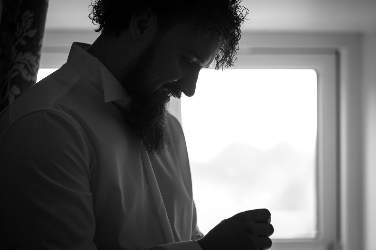 Black and white portrait of a bearded man in a white shirt looking down near a bright window.