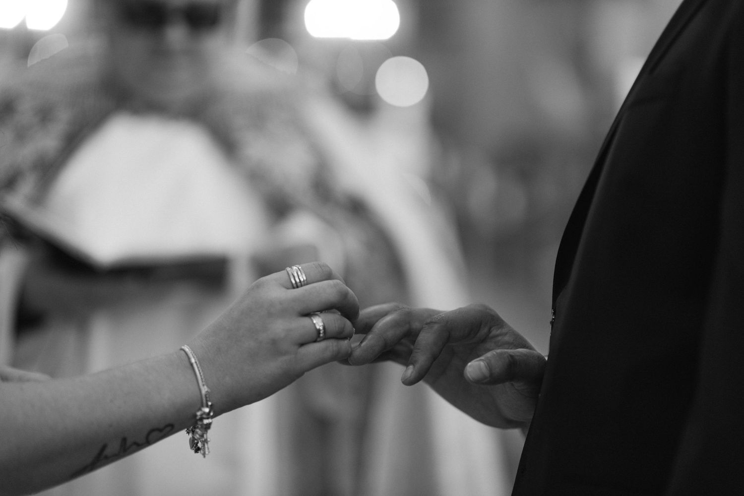 Black and white photo of a bride placing a wedding ring on a groom's finger during a ceremony with an officiant blurred behind.