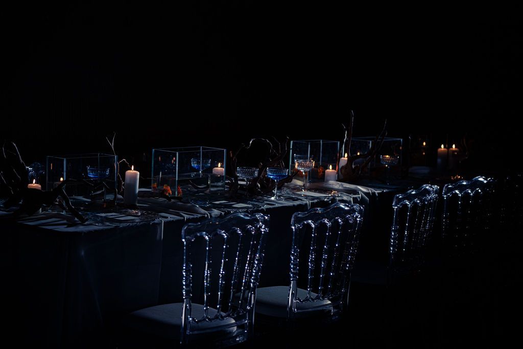 Elegant candlelit banquet table with clear acrylic ghost chairs, glowing candles and floral centerpieces in a dark, moody event venue.