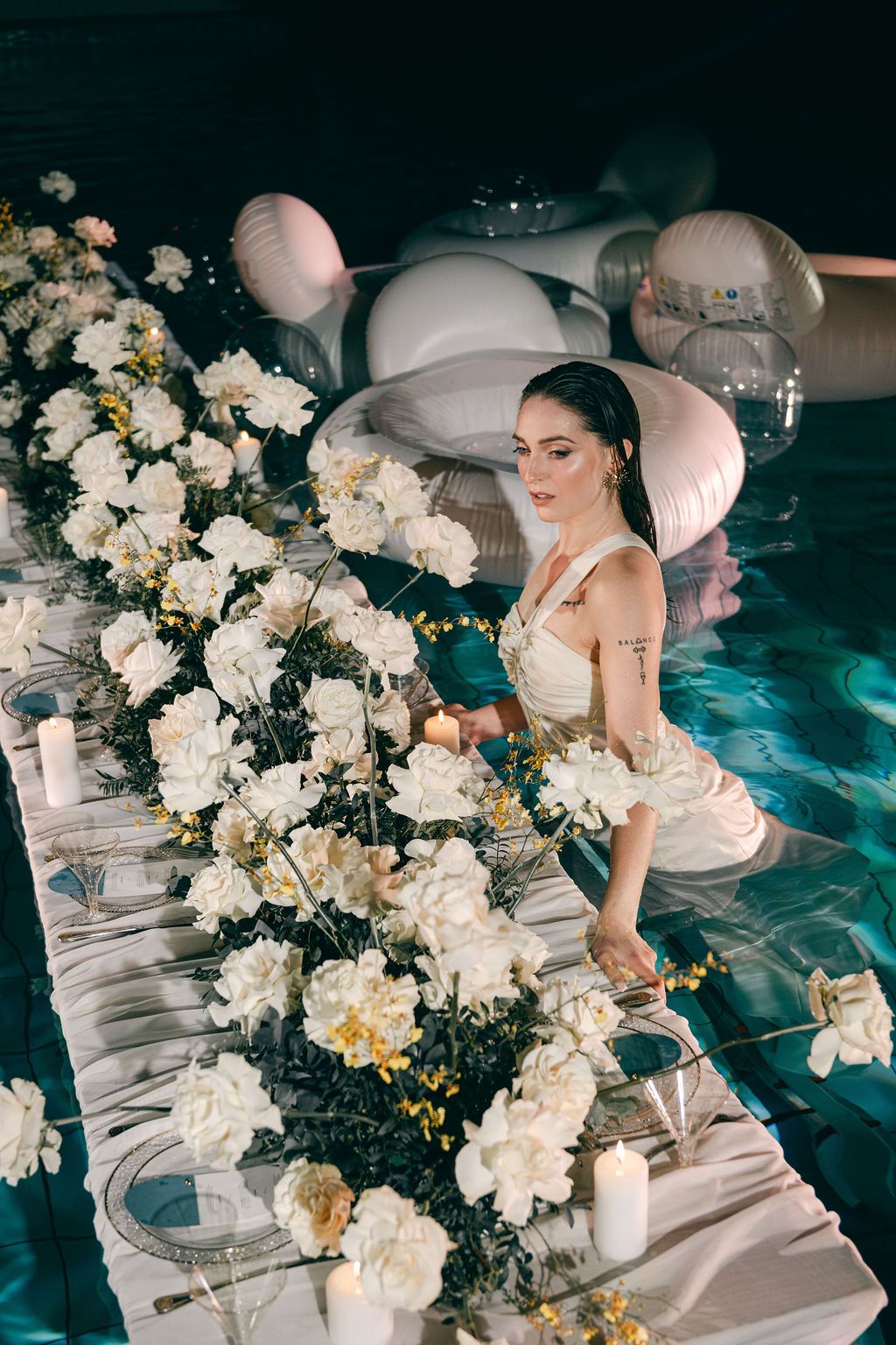Woman in a white dress stands in a pool beside a floating table of white roses, pillar candles and inflatable decor at night.