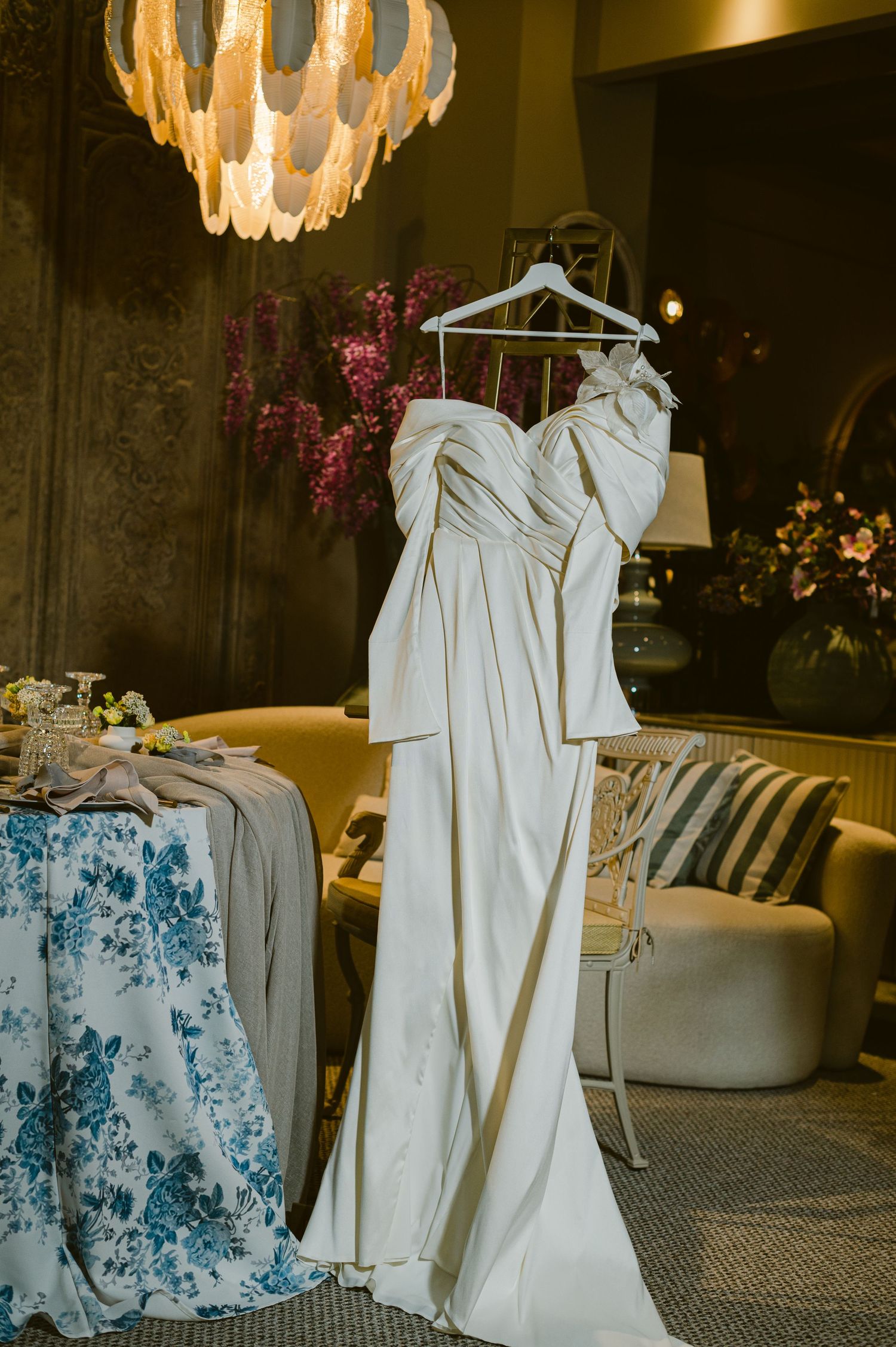 Two elegant white wedding dresses hang beside a blue floral dress in a luxurious room with chandelier lighting.