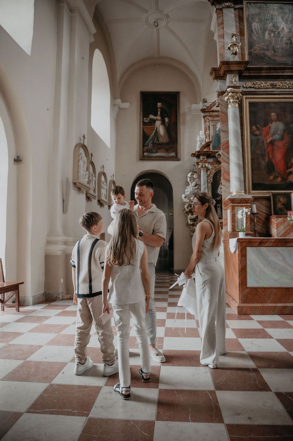 Family in white attire gathered inside an ornate baroque church with checkered marble floors and religious paintings.