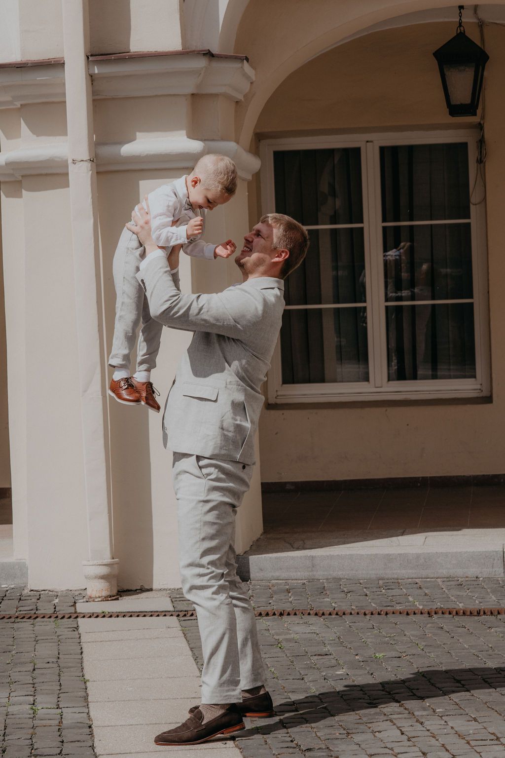 A smiling father in a gray suit lifts his happy toddler son high in the air outdoors.