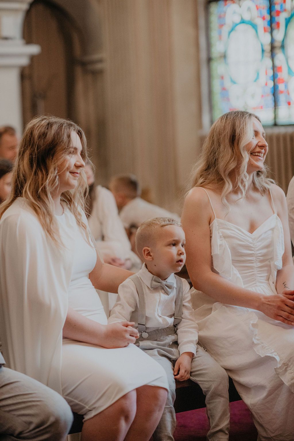 Two women in white dresses and a young boy in a bow tie seated in a church pew during a wedding ceremony.