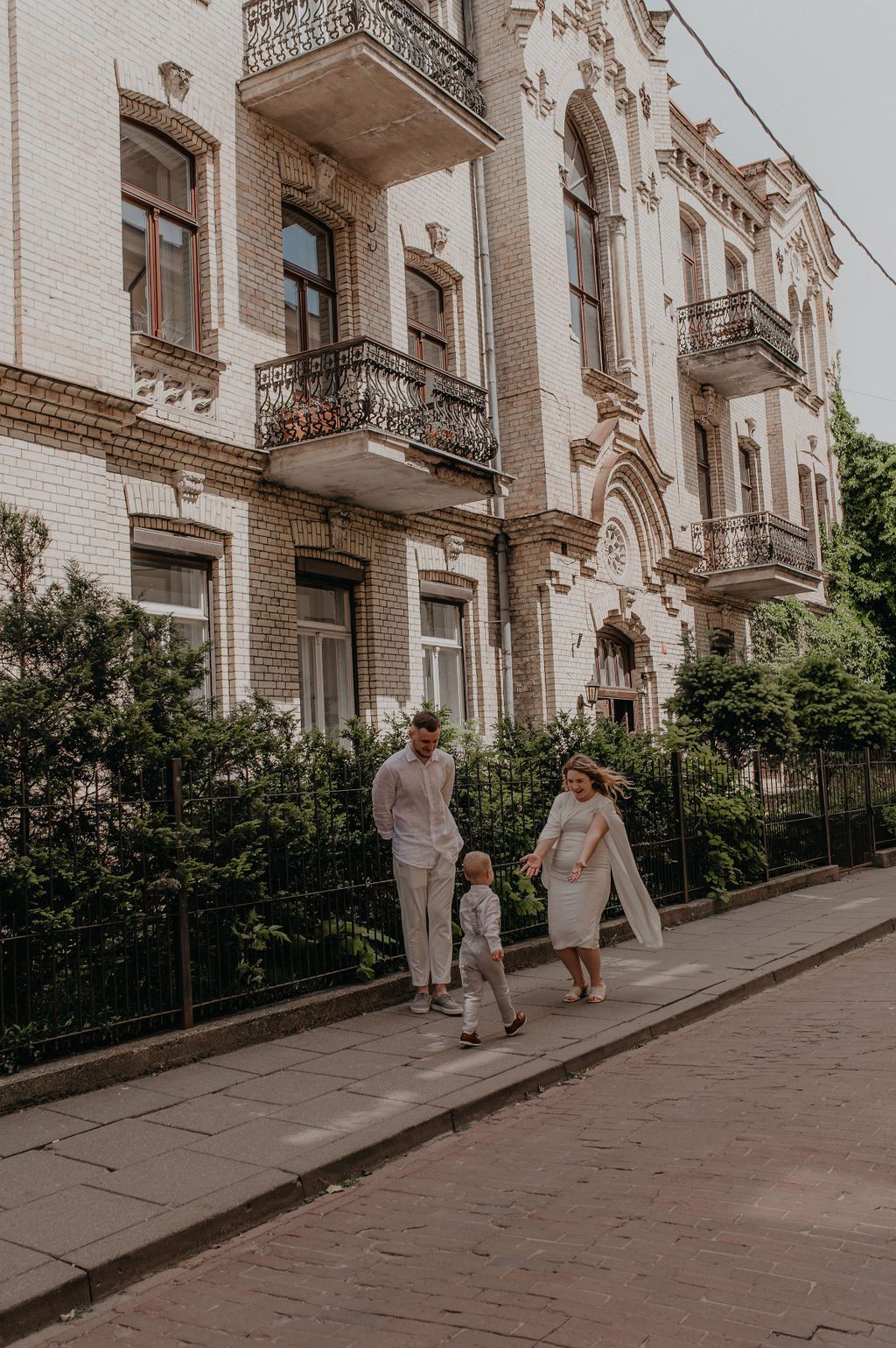 A family of three strolls along a sidewalk in front of an ornate white building with balconies.