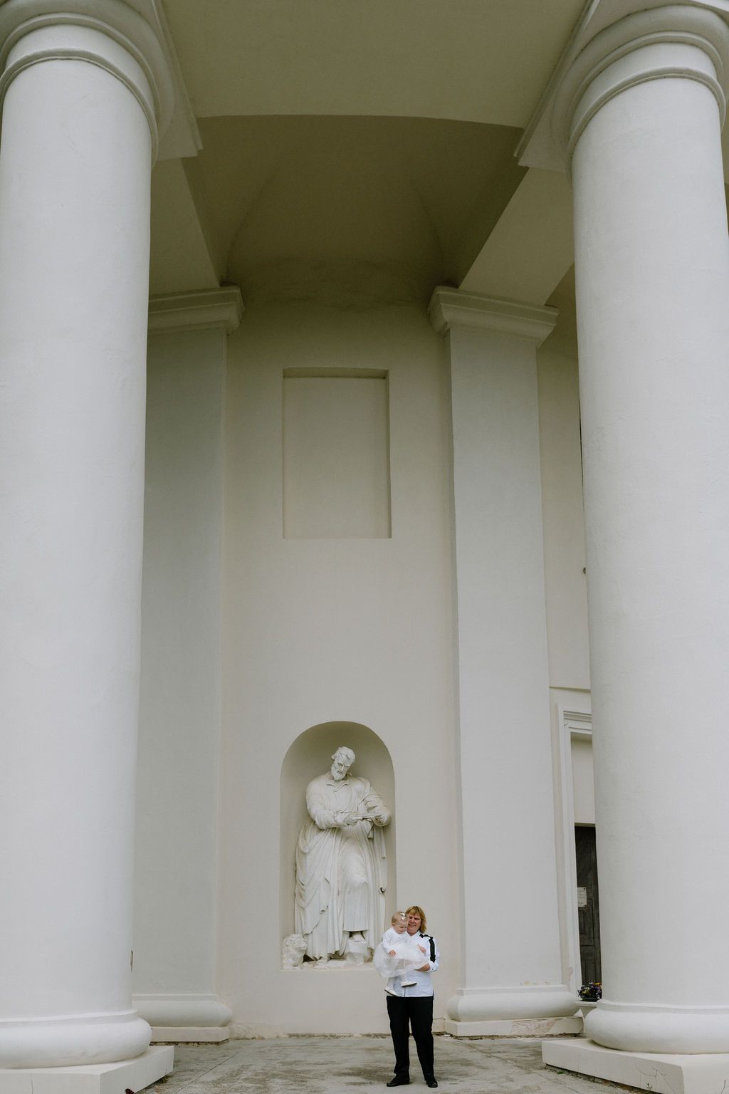 Woman stands between white columns before a classical building with a statue in an arched niche.