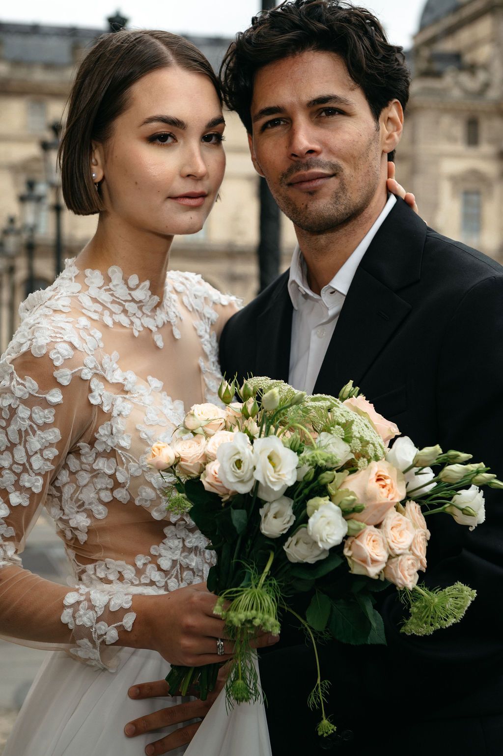 Elegant bride and groom posing with a white and blush rose bouquet, bride wearing lace wedding gown, Paris backdrop.
