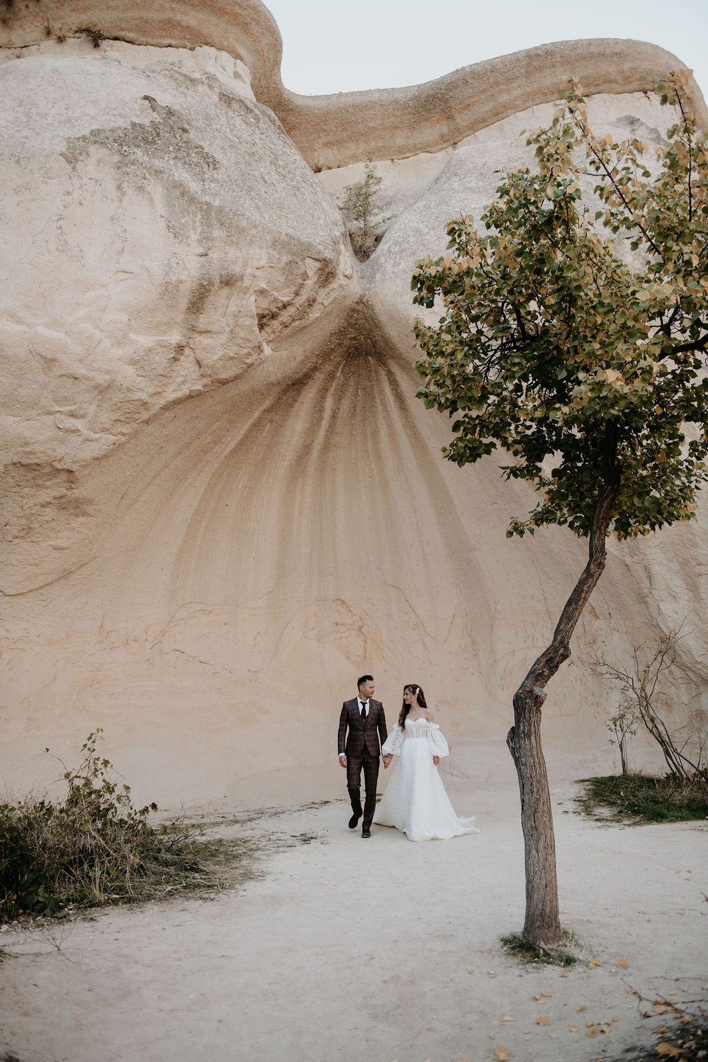 Bride and groom walk hand-in-hand near a dramatic sandstone rock formation and tree in Cappadocia, Turkey.