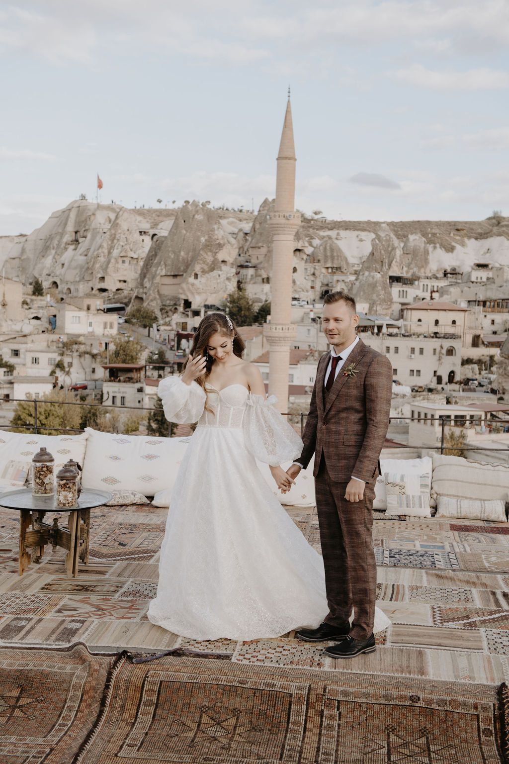 Bride and groom hold hands on a rooftop in Cappadocia, Turkey, with cave dwellings and a minaret in the background.