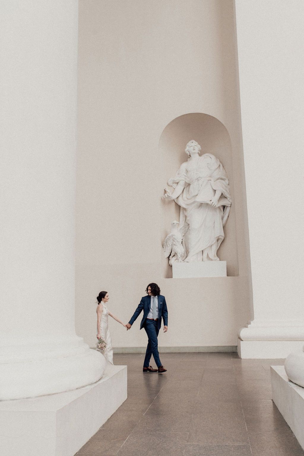 Couple holds hands walking past a classical white marble statue in an elegant neoclassical hall.