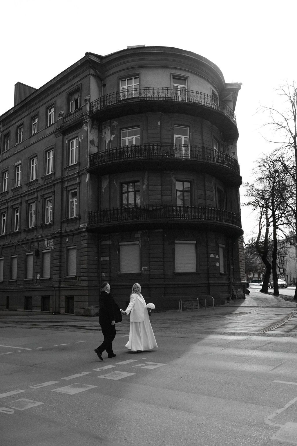 Black and white photo of a bride and groom crossing a street in front of a classic European corner building.