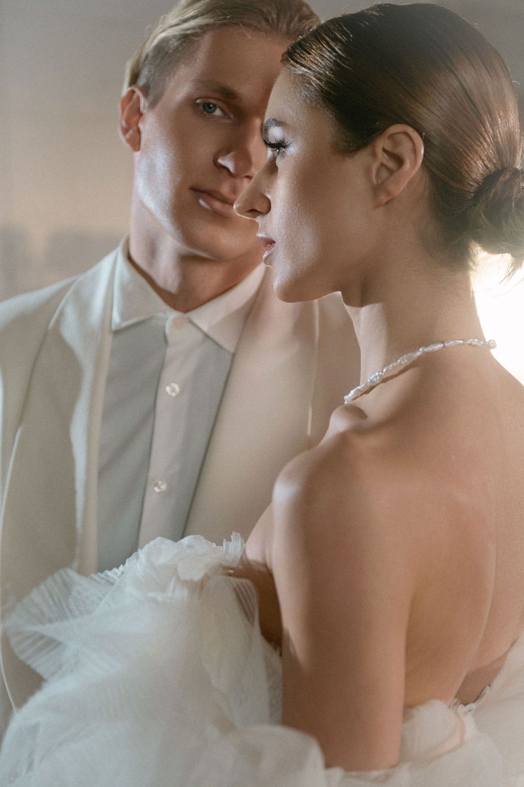 Elegant bride and groom in white wedding attire share an intimate moment, soft backlit photography.