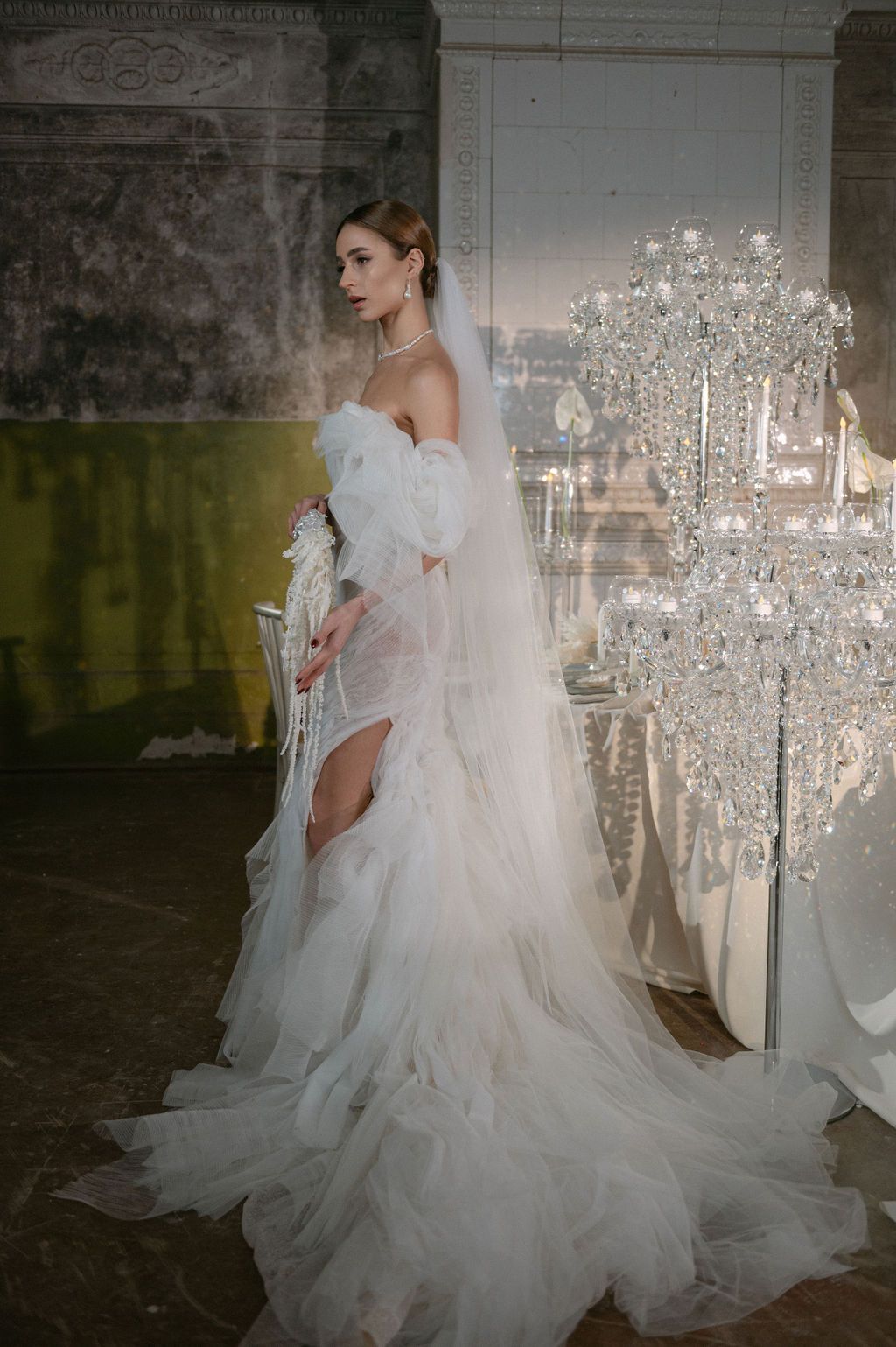 Bride in sheer white tulle wedding gown with off-shoulder sleeves and veil poses near crystal chandelier.