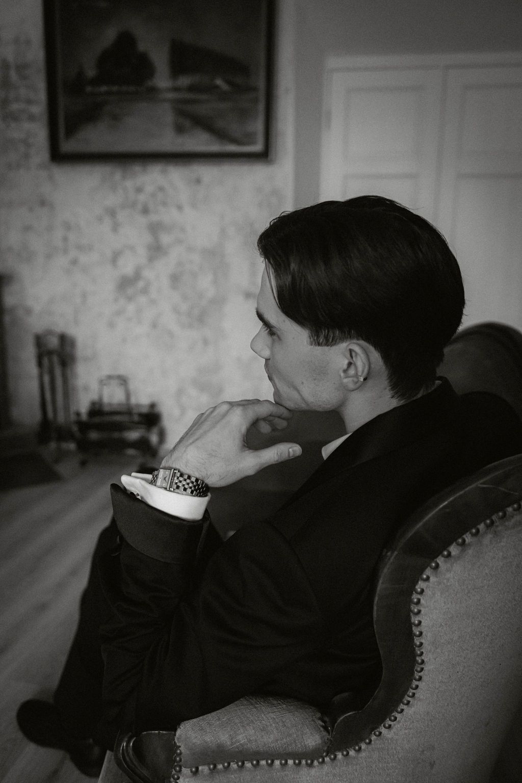Thoughtful man in black suit sitting in vintage armchair in black and white contemplative portrait.