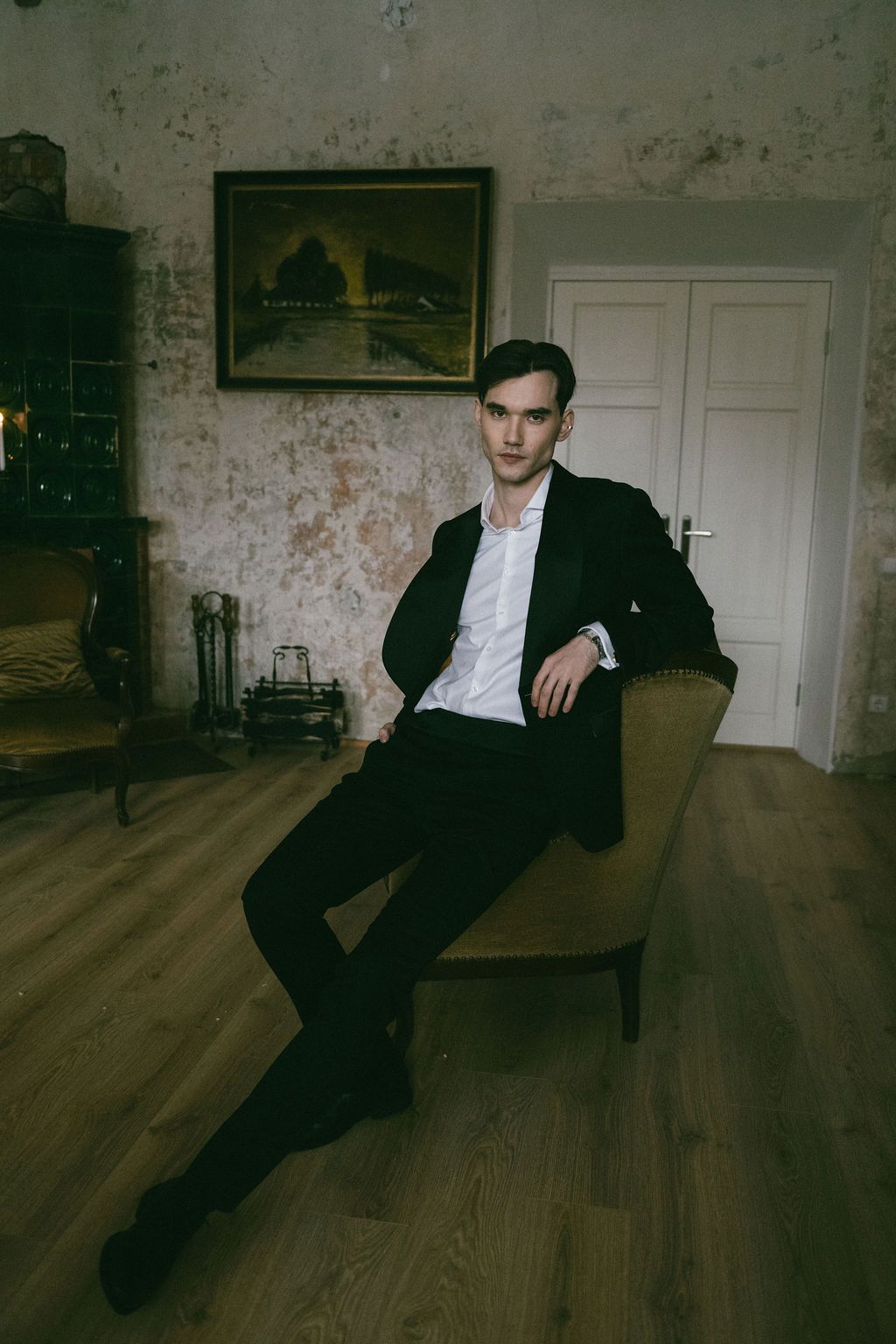 Young man in black suit sitting elegantly in vintage armchair inside rustic room with wooden floors and classic painting.