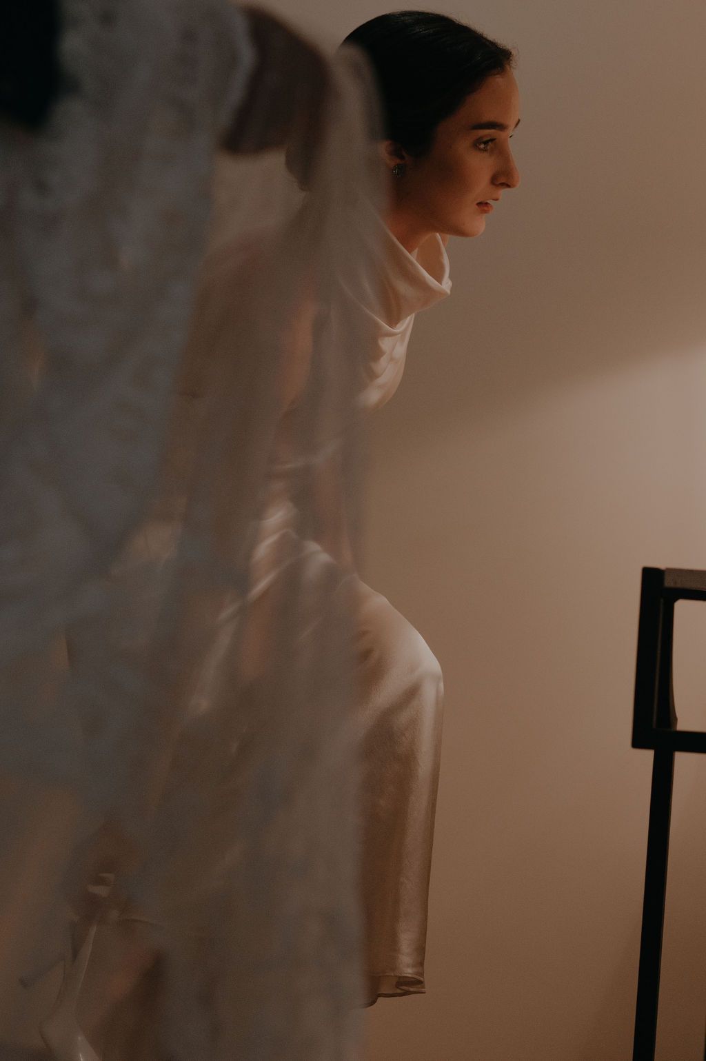 Bride in elegant white satin high-neck wedding dress with veil, standing in soft warm light.