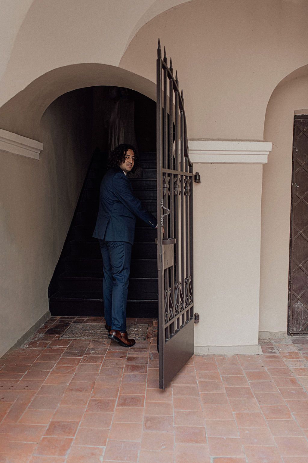 Man in navy blue suit opening ornate iron gate at arched stone entrance with terracotta tile floor.