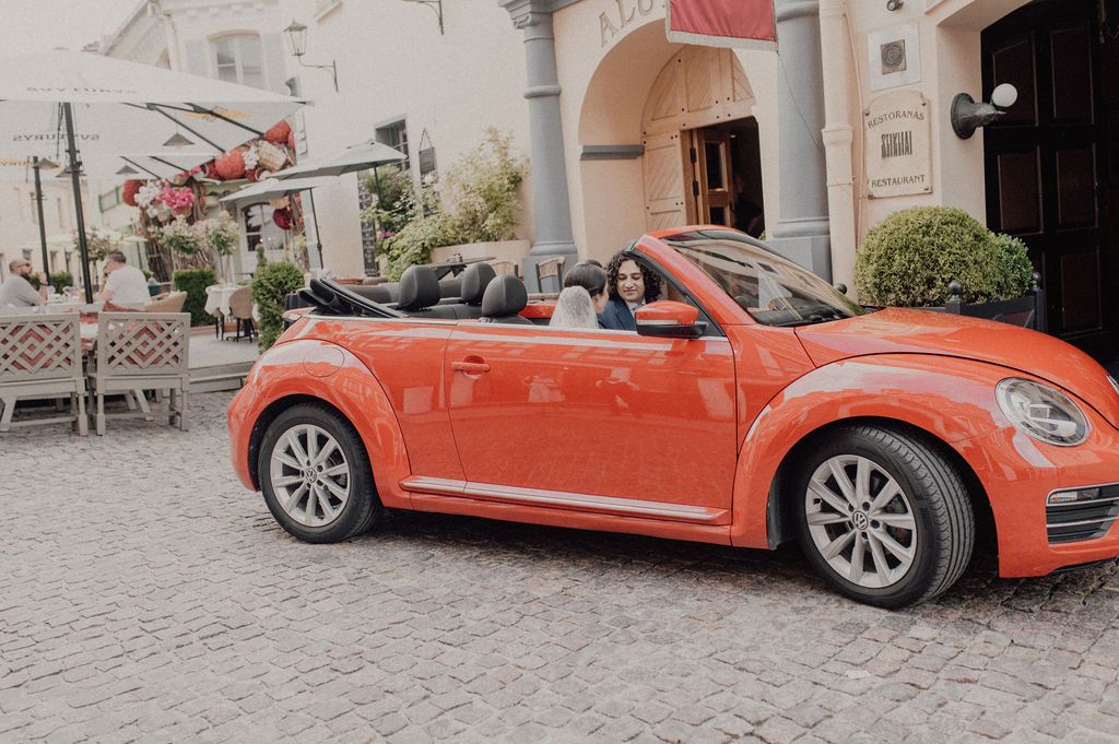 Orange VW Beetle convertible parked on cobblestone street near outdoor cafe.
