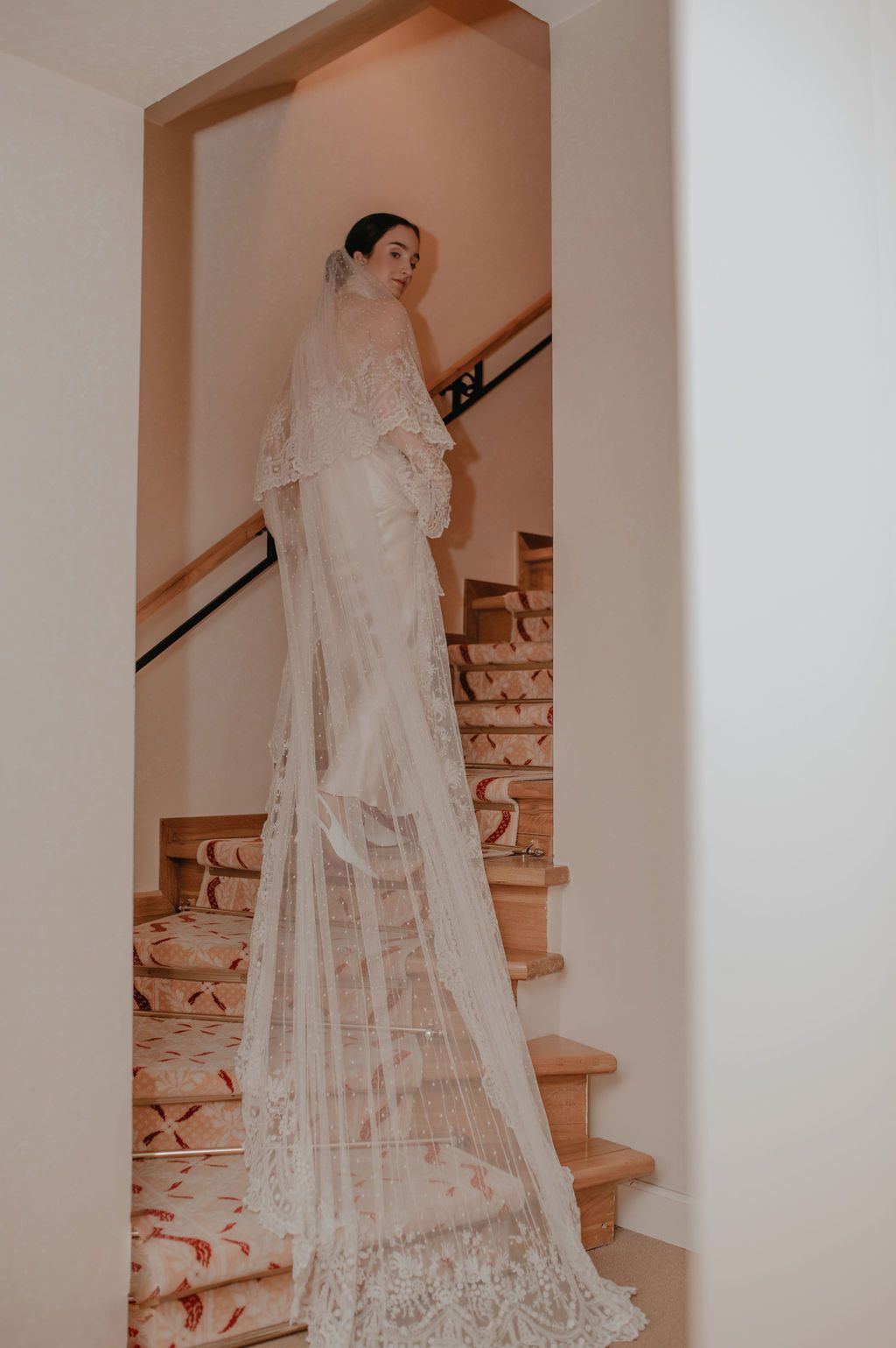Bride in lace wedding gown with long cathedral veil descending ornate staircase.