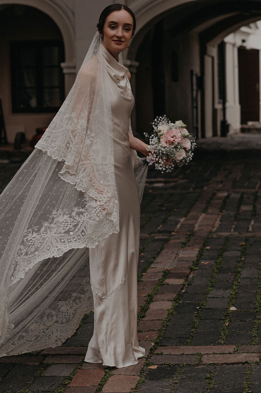 Elegant bride in ivory silk slip dress and lace cathedral veil holds pink floral bouquet in cobblestone courtyard.