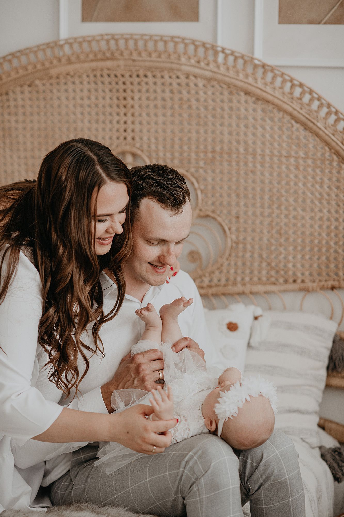 Parents in white shirts share tender moments with their newborn baby on a rattan chair with cushions.