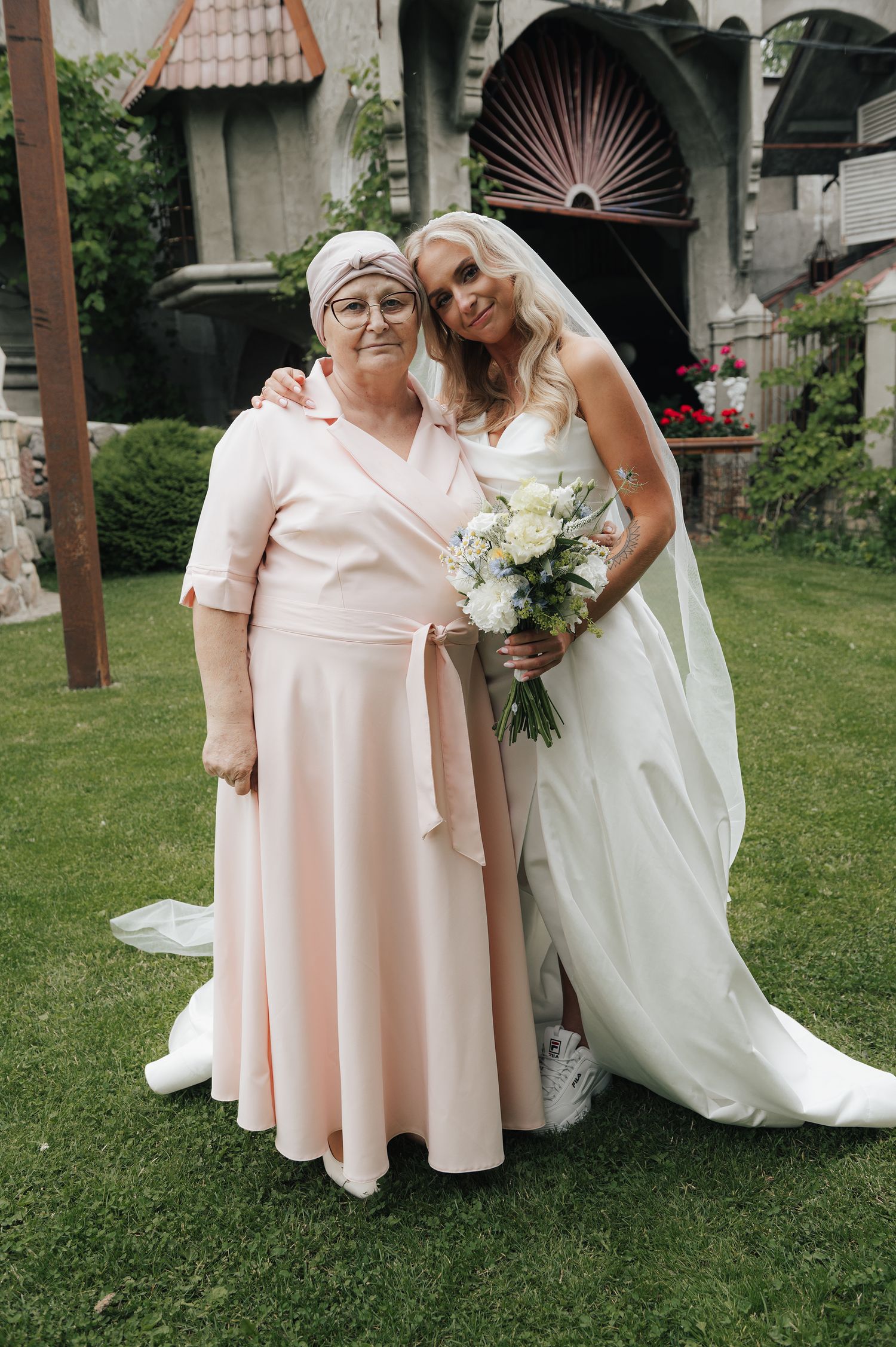 A bride in a white wedding dress kneels beside someone wearing a light pink dress on a grassy lawn.