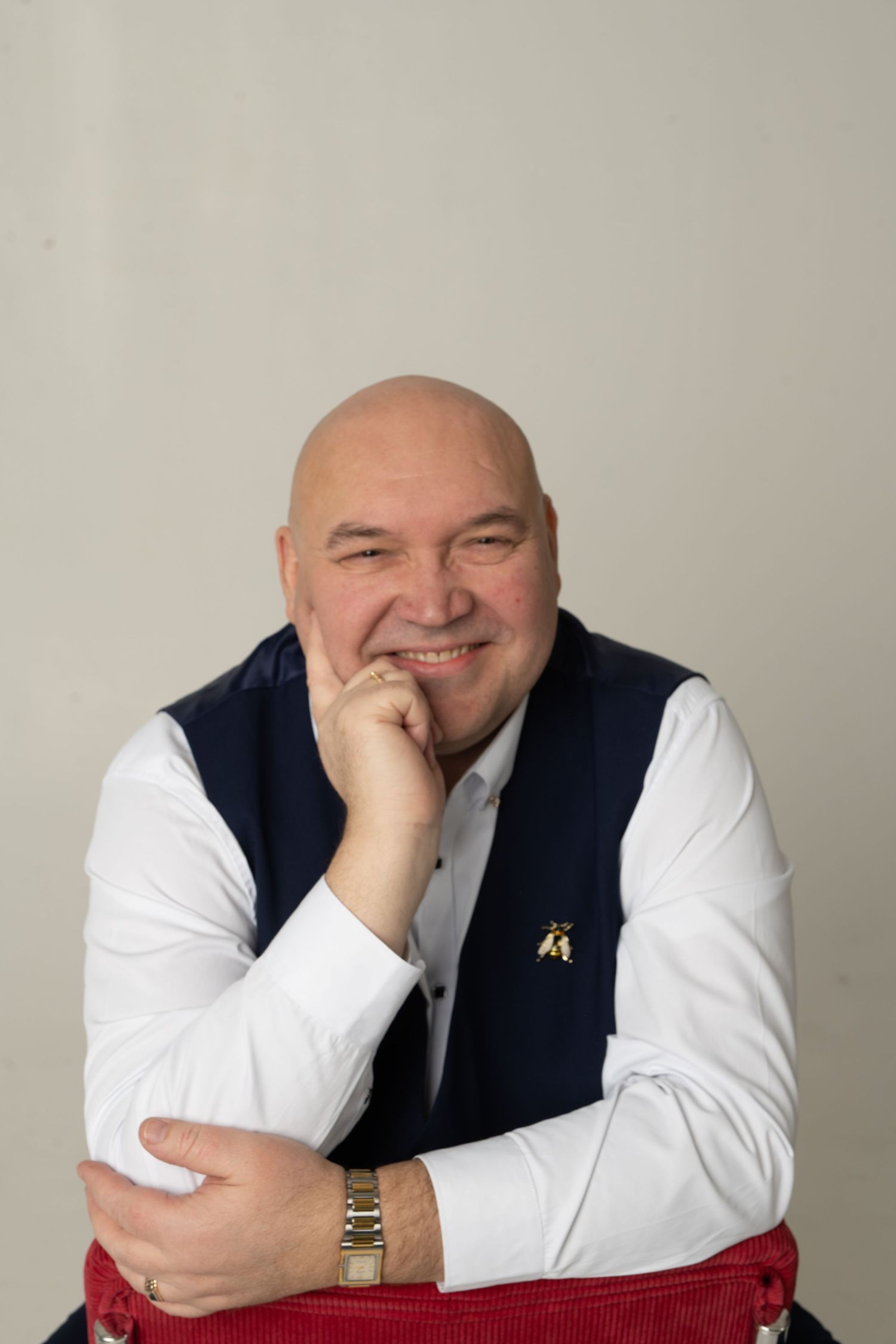 Smiling bald man in white shirt and navy vest, posing with chin resting on hand in a professional studio portrait.