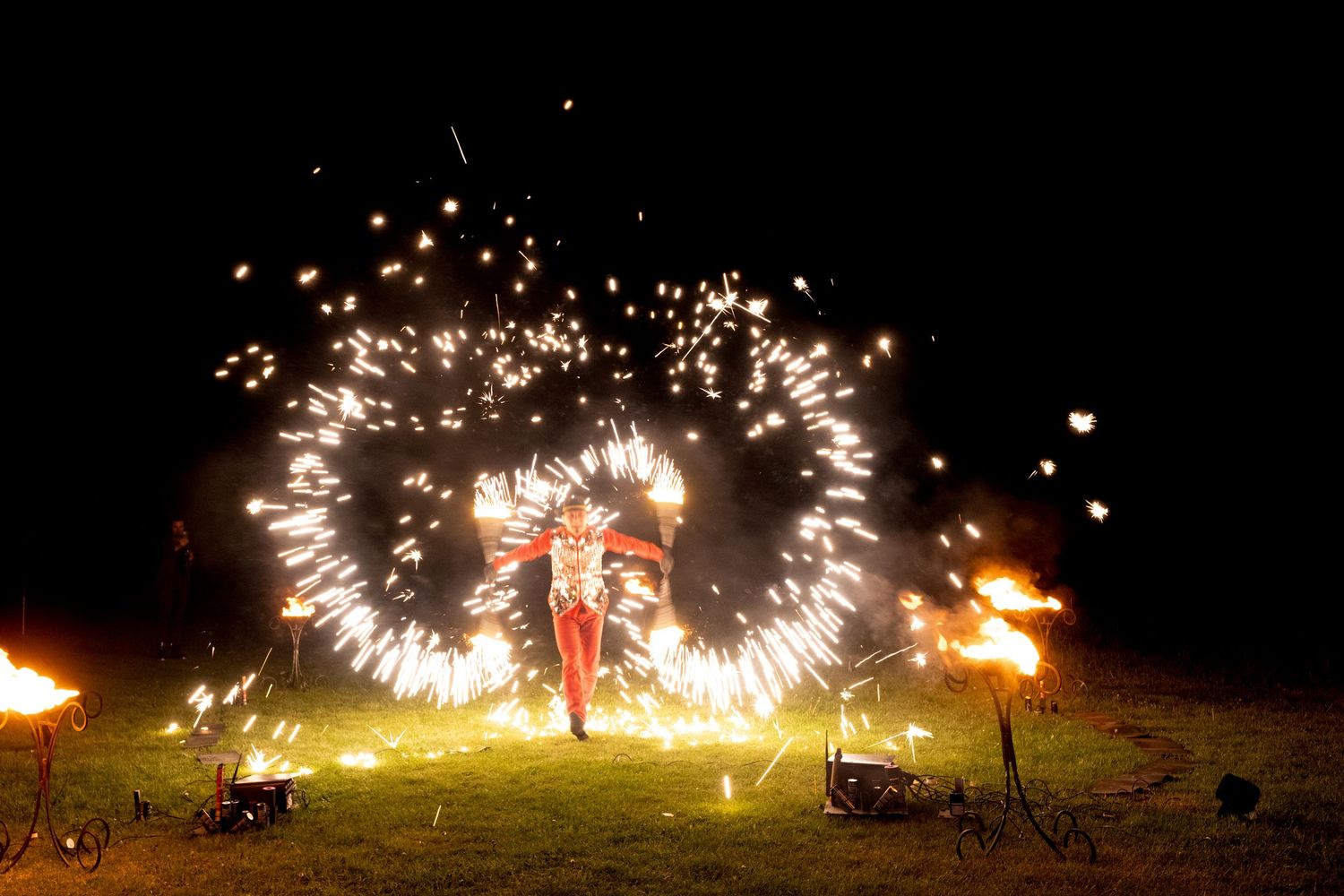 Fire performer in red costume spinning glowing sparkler rings at night, surrounded by burning torches on a grassy field.